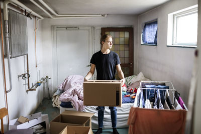 Full length of thoughtful man carrying cardboard box while sanding in bedroom