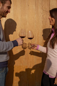 Couple stand with glasses of wine opposite each other on a wooden background with beautiful shadows