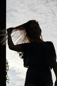 Low angle view of young woman holding long blond hair against cloudy sky