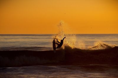 Silhouette person surfing in sea against orange sky during sunset