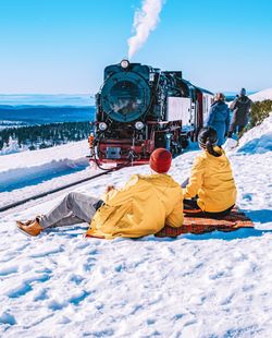 Couple sitting on snow covered land at railroad station against clear blue sky