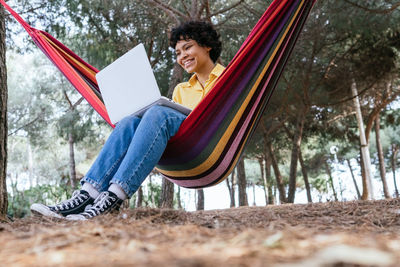 Ground level of smiling teen female chilling in hammock in park and browsing laptop