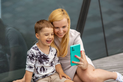 Happy blond woman and little boy sitting on terrace and making selfie on smartphone. mother and son