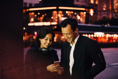 Smiling man and woman using smart phone while standing in city at night