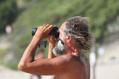 Side view of shirtless senior man looking through binoculars at beach