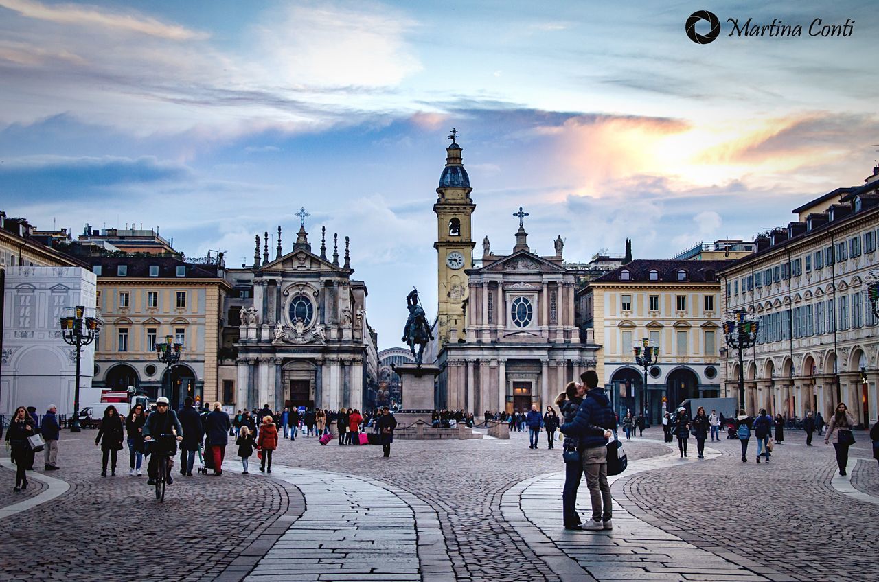 building exterior, large group of people, architecture, travel destinations, travel, city, sky, outdoors, town square, city life, built structure, tourism, cloud - sky, people, ice rink, clock tower, day, clock, adults only, astronomical clock, adult