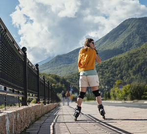 Rear view of young woman in protective equipment roller skating along embankment against mountains 