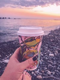 Low section of woman holding disposable cup at beach during sunset