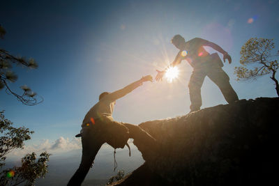 Low angle view of young man assisting friend in climbing rock against sky during sunny day