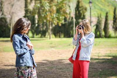 Woman photographing smiling friend in park during sunny day