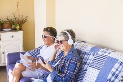 Smiling senior woman listening music through headphones by grandson sitting on sofa at home