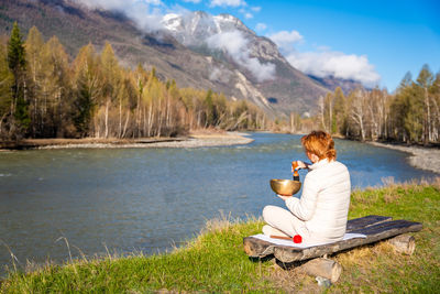 Side view of woman sitting on rock by lake against mountain