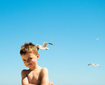 Low angle view of shirtless boy against seagulls flying in clear blue sky