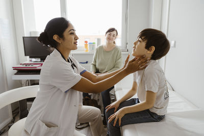 Female pediatrician examining neck of boy in examination room at hospital