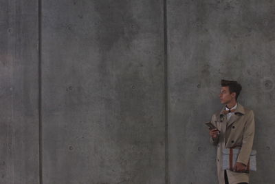 Young man holding mobile phone while standing against wall