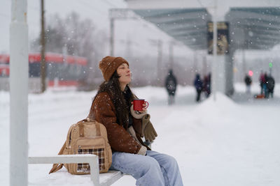 Asian girl sitting on bench drinking hot tea while waiting for arrival of train on snowy winter day