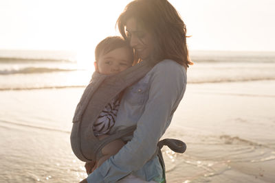 Mother carrying son in baby carrier while standing at beach