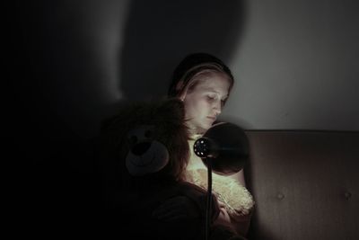 Young woman sitting with stuffed toy by illuminated lamp on sofa at home