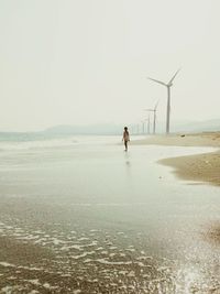 Girl standing on shore at beach with wind turbines against sky
