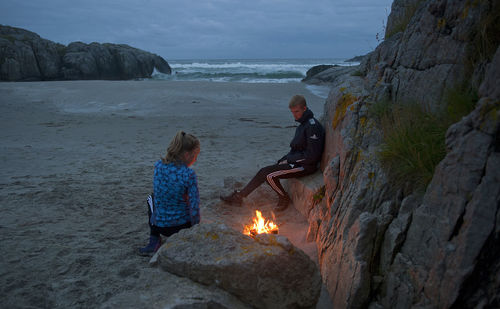 Siblings sitting by campfire at beach during dusk