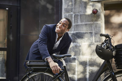 Smiling young male commuter locking electric bicycle while looking away against building in city