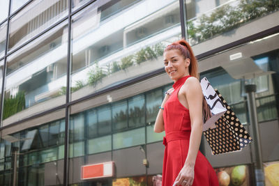 Low angle portrait of young woman holding shopping bag while standing against building