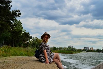 Portrait of smiling young woman sitting by tree against sky