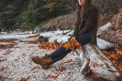 Low section of woman holding leaves while sitting on tree trunk