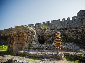 Boy looking away while walking against built structure and clear sky