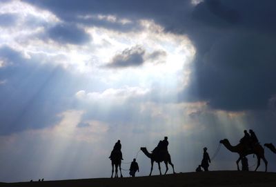 Silhouette people and camels against cloudy sky