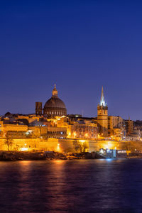 Illuminated buildings by river against clear sky at night