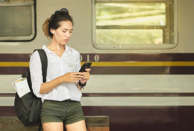 Young woman using mobile phone while standing against train at railroad platform