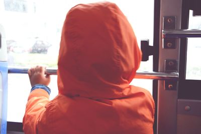 Rear view of kid in orange hooded jacket standing by bus door
