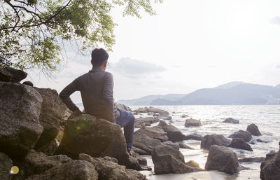 Rear view of man sitting on rock by sea against sky