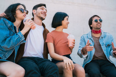 Smiling friends holding juices sitting against wall outdoors