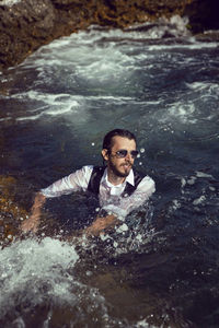 Man with a beard and sunglasses in clothes a vest and a white shirt swims in the sea among the rocks