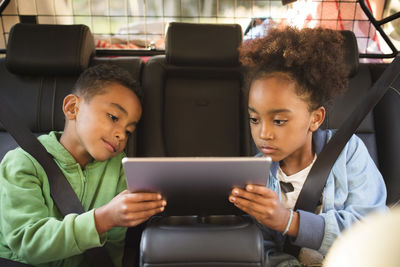 Siblings sharing digital tablet while sitting in electric car