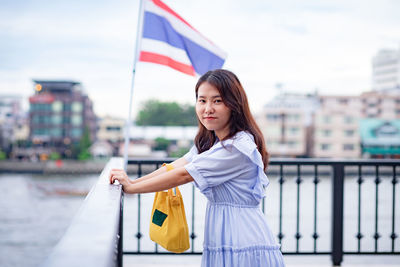 Portrait of woman standing by railing against flag and buildings in city