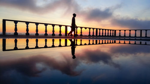 Symmetric view of silhouette man walking by swimming pool against sky during sunset