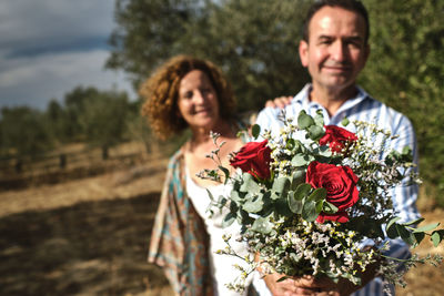 Cheerful middle aged couple in casual wear standing together with tender bouquet of flowers while enjoying stroll in summer garden on sunny day