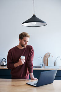 Man holding coffee cup while looking at laptop at home