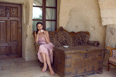 Woman sits on an old sofa at the entrance to the house in the rock and drinks tea