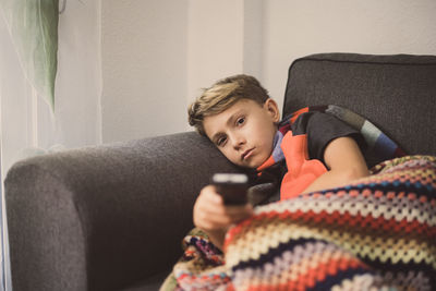 Portrait of ill boy holding remote control while relaxing on sofa at home