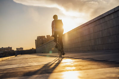 Young man with backpack riding bike on promenade at sunset