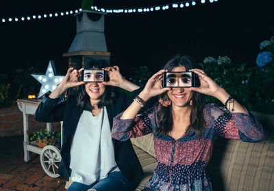 Portrait of young friends holding smart phones on eyes while on sofa during party