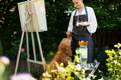 Artist working on her painting outdoors in her garden with golden retriever keeping her company. 