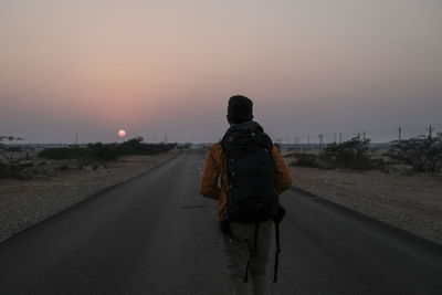 Rear view of tourist with backpack walking on country road against sky during sunset