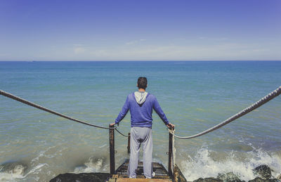 Rear view of man standing on steps by blue sea against sky