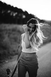 Content young female in casual wear and summer hat holding cruiser skateboard and looking at camera while standing on empty asphalt road in rural area at sunset