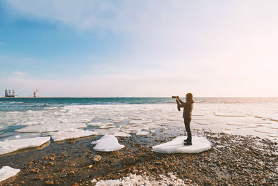 Full length of man photographing through camera while standing on ice against sea at beach